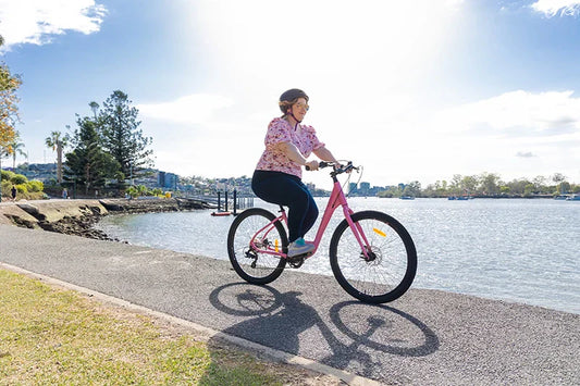 Electric bikes for urban commuters. Pink ebike from Bicycle Centre ridden along the river with buildings in the background. Lady commuting to the office on her step-thru electric bike.